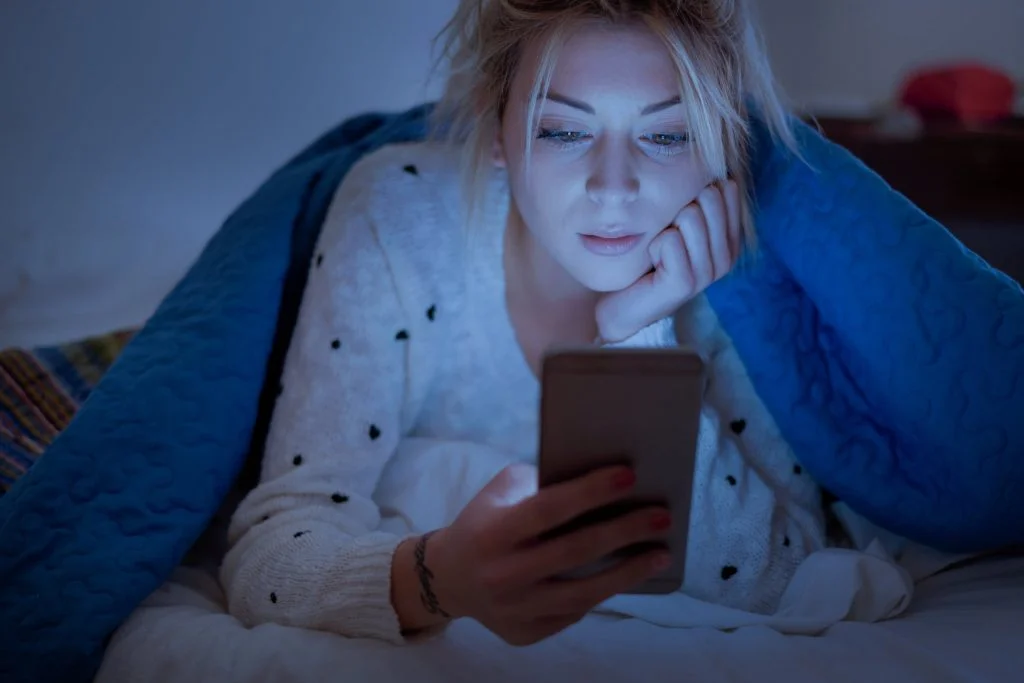 woman holding mobile phone while laying on bed at night. exposure to blue light